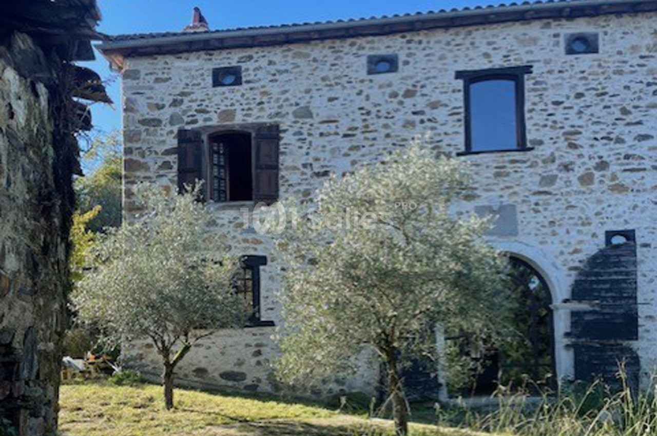 Façade en pierre d'une maison ancienne avec volets en bois, entourée d'arbres et d'herbe.