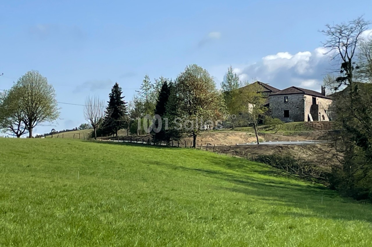 Vue d'une prairie verdoyante avec des arbres et une maison en pierre sous un ciel bleu dégagé.