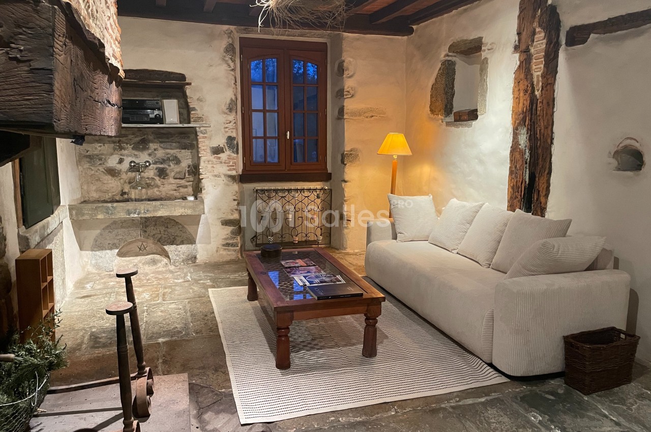 Salon rustique avec murs en pierre, poutres apparentes, canapé blanc, table basse en bois et lampe allumée.