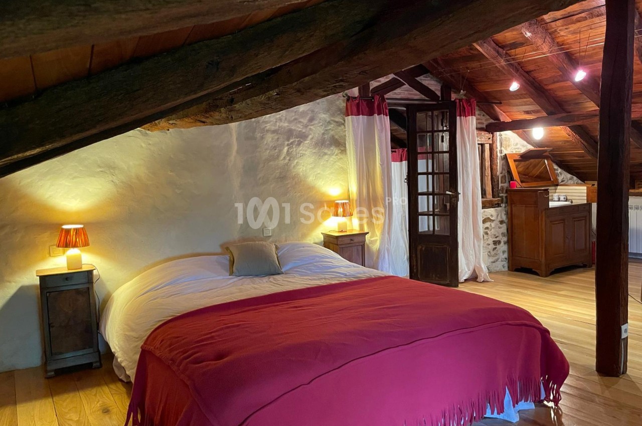 Chambre mansardée avec lit double, couvre-lit rouge, murs en pierre et poutres apparentes, éclairée par des lampes de chevet.