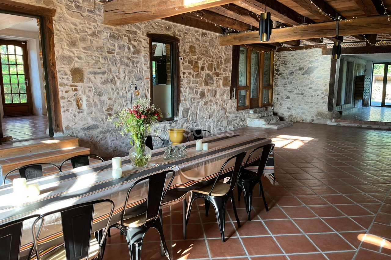 Salle lumineuse avec murs en pierre, poutres apparentes, grande table décorée et carrelage en terre cuite.