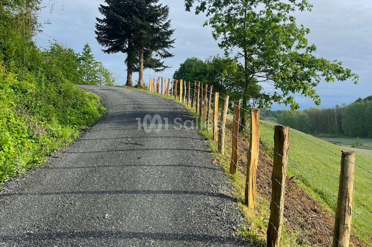 Chemin de gravier bordé de clôtures en bois, serpentant à travers une campagne verdoyante sous un ciel nuageux.