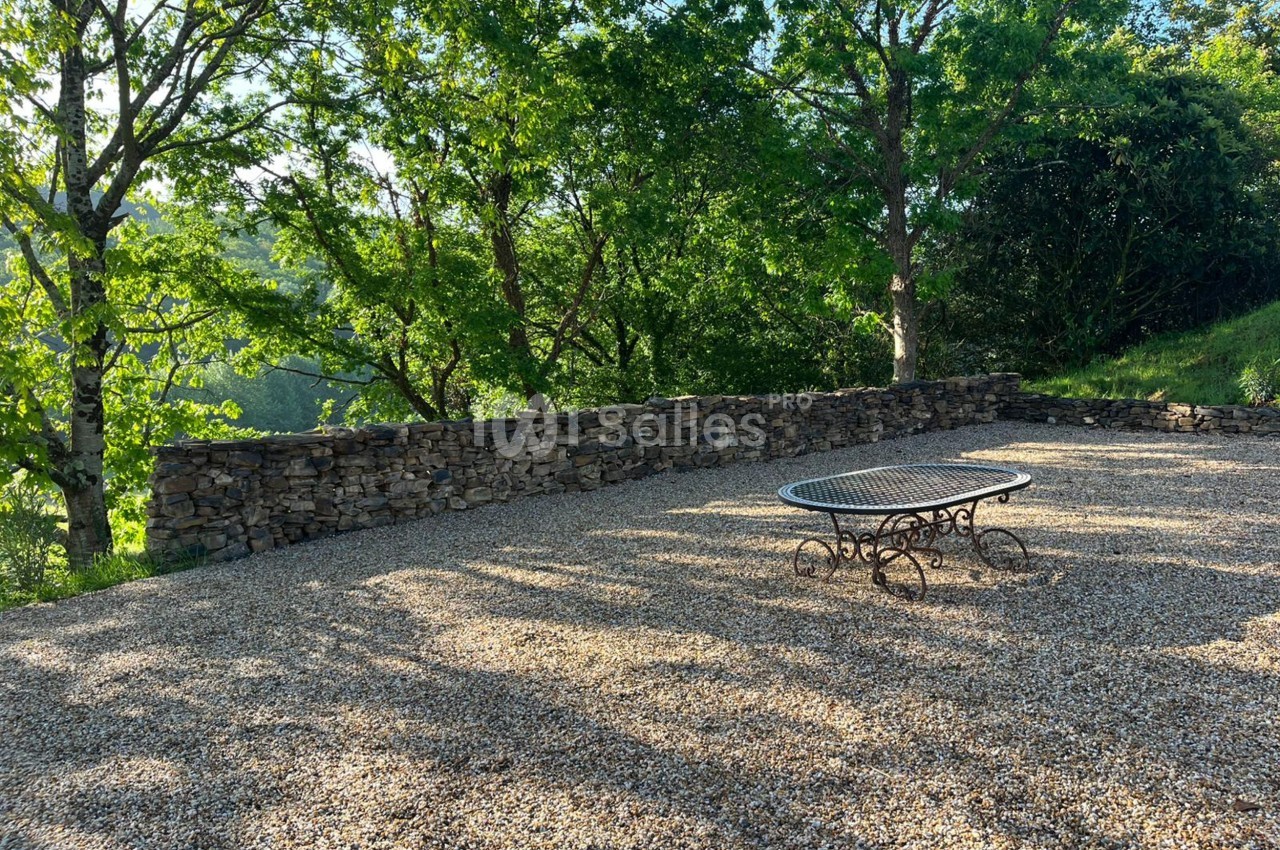 Terrasse en gravier avec une table en fer forgé, entourée d'arbres et d'un muret en pierre sèche.