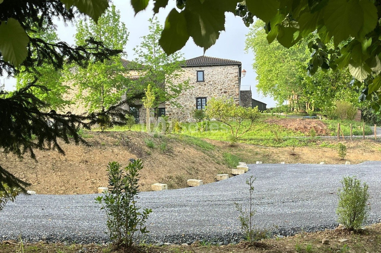 Maison en pierre entourée de verdure, avec une allée en gravier et des arbres en premier plan.