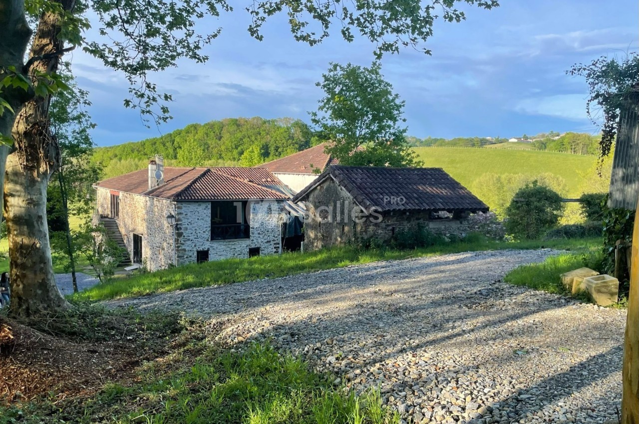 Chemin de gravier menant à des bâtiments en pierre entourés de verdure et de collines sous un ciel dégagé.