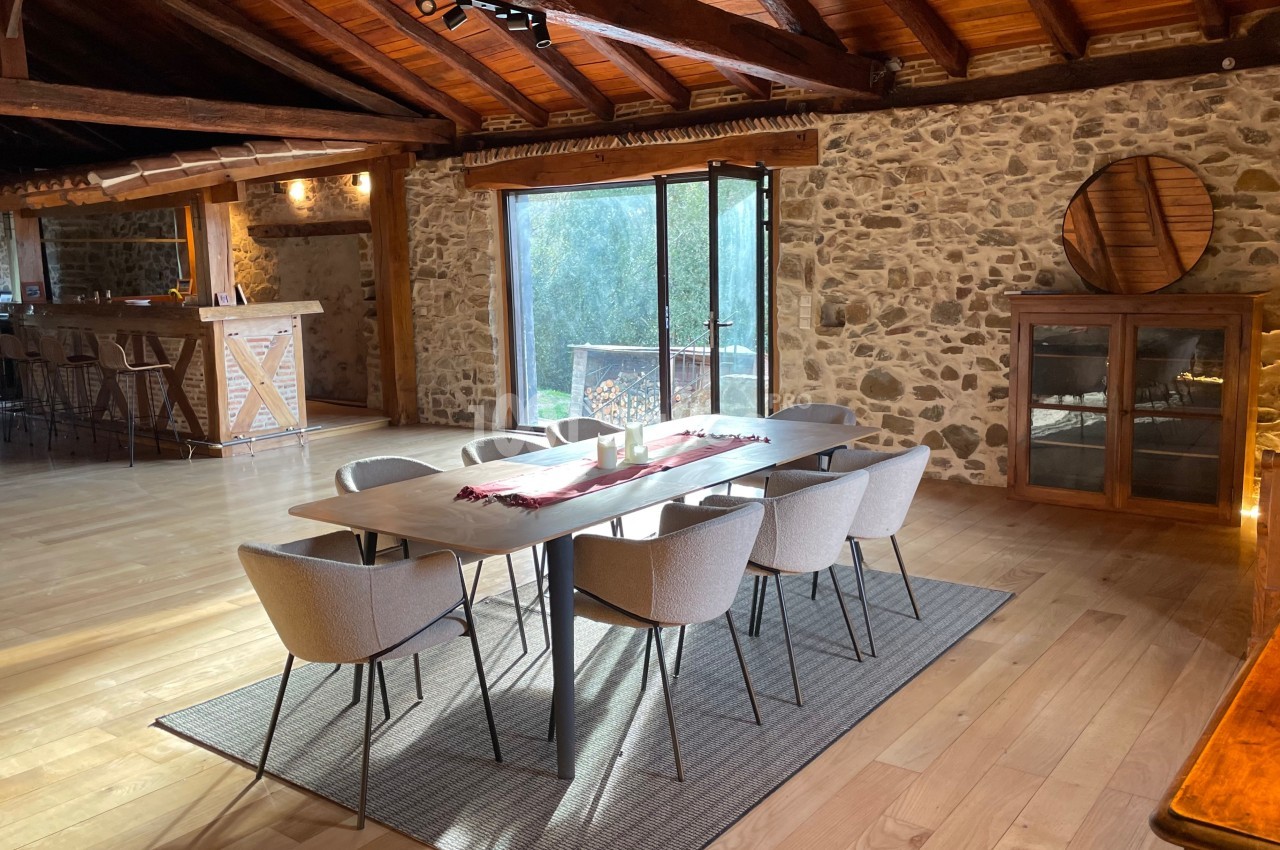 Salle à manger lumineuse avec table moderne, chaises rembourrées, murs en pierre et plafond en bois apparent.