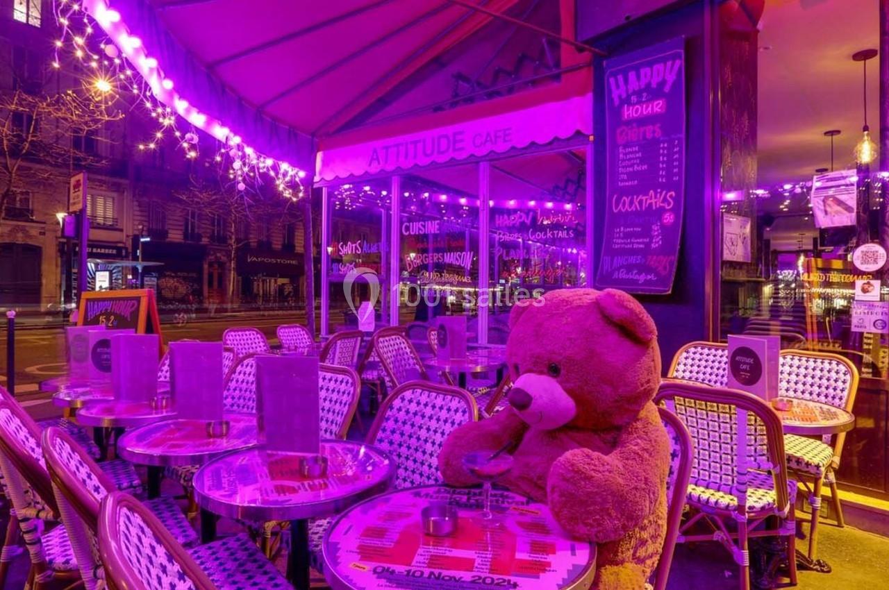 Terrasse d'un café illuminée par des lumières violettes, avec un grand ours en peluche assis à une table.