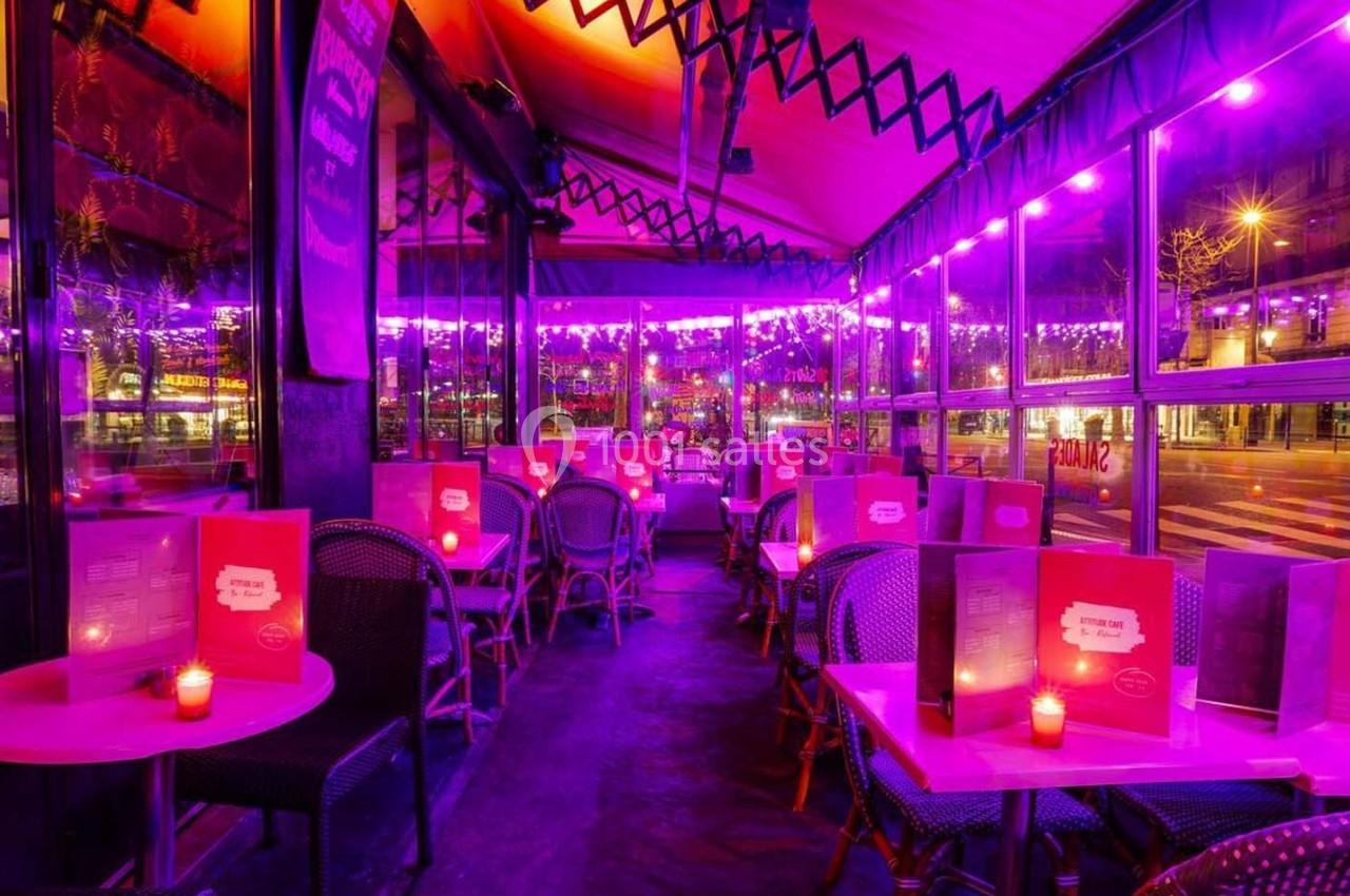 Salle de restaurant avec tables rondes, menus roses et éclairage violet, donnant sur une rue illuminée.