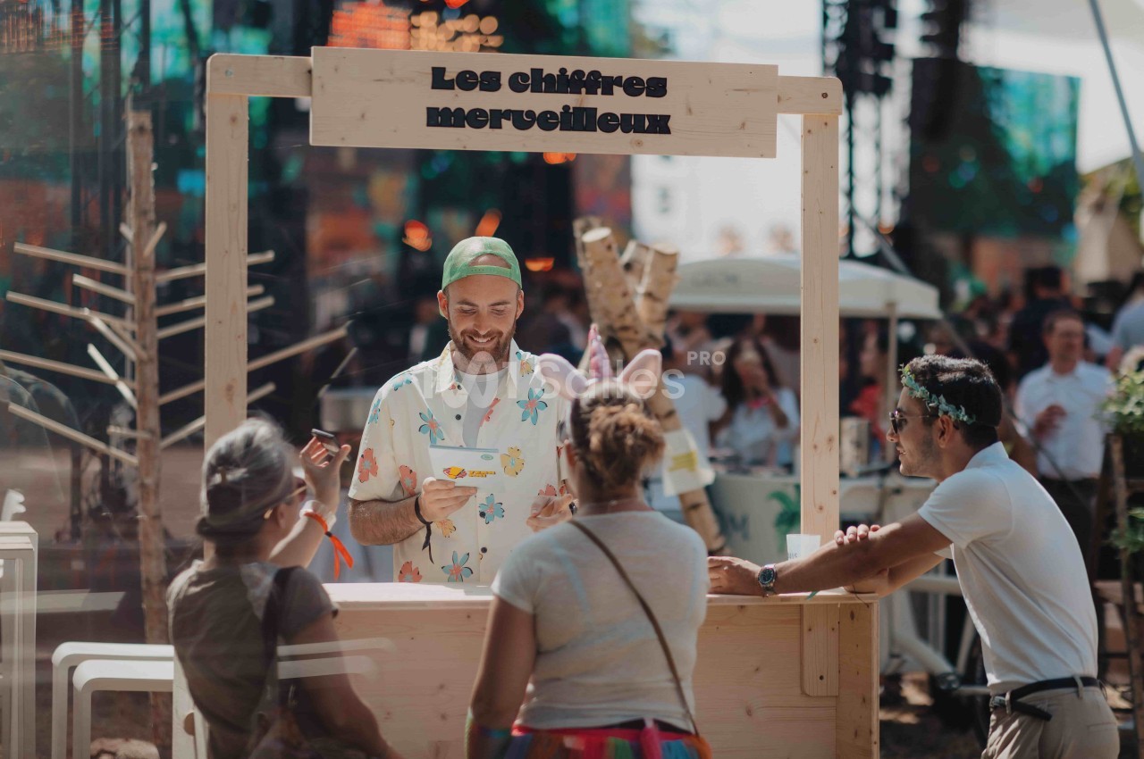 Un homme souriant derrière un stand en bois interagit avec trois personnes dans un environnement festif en extérieur.