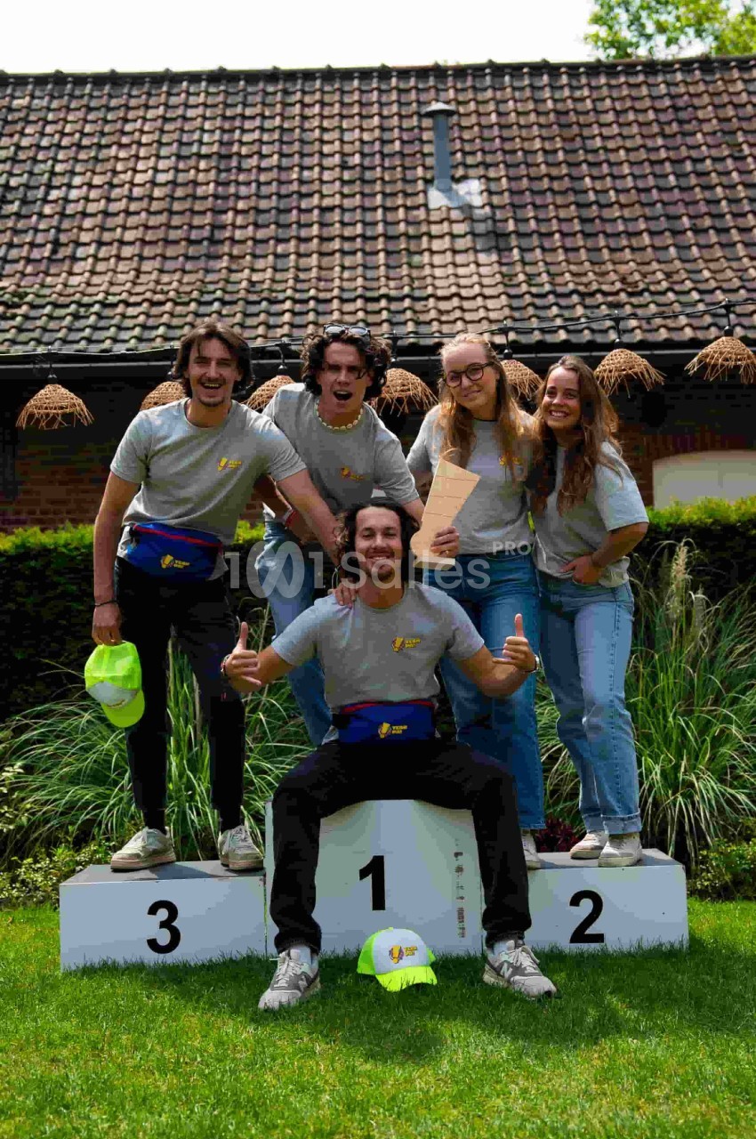 Un groupe de cinq personnes souriantes posant sur un podium dans un jardin devant un bâtiment en briques.
