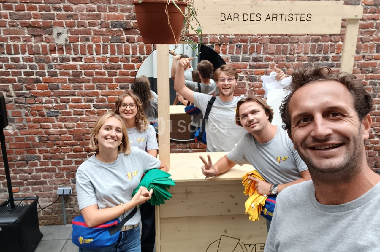 Des personnes souriantes posent devant un stand en bois marqué ’Bar des artistes’, dans une cour en briques.