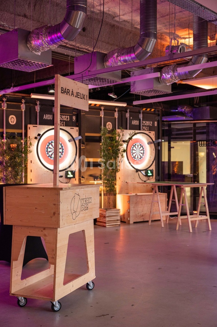 Espace intérieur avec cibles de fléchettes lumineuses, bar en bois et ambiance industrielle.