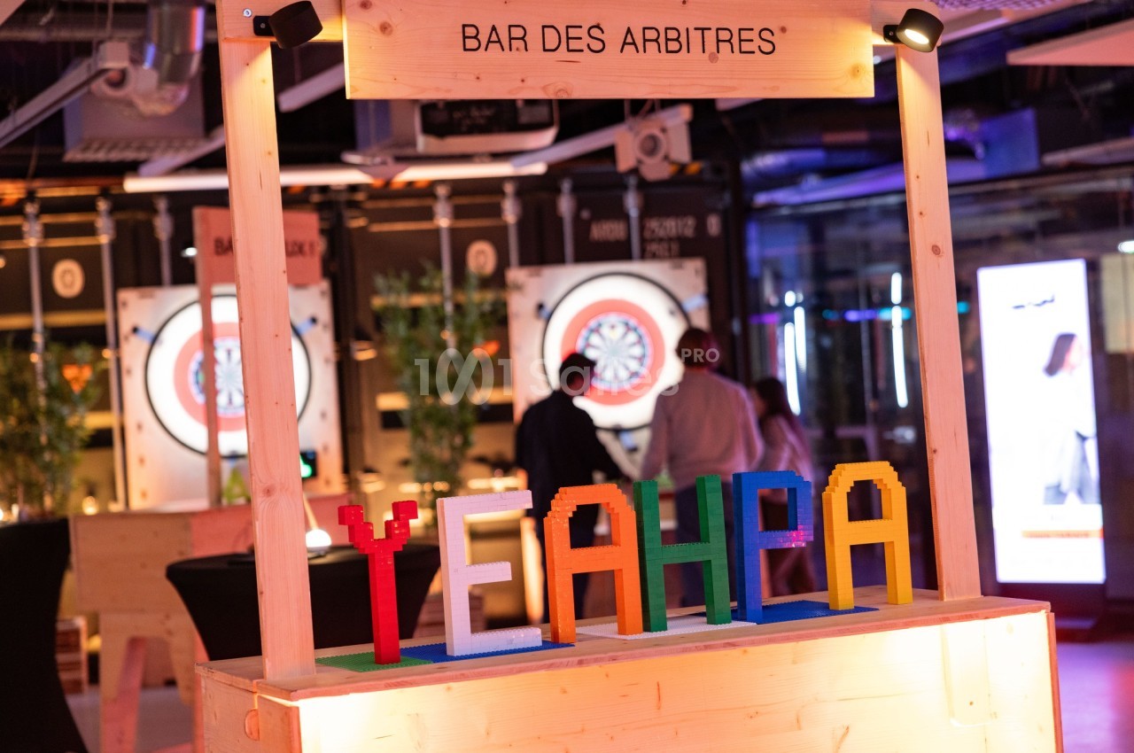 Stand en bois avec l'inscription ’Bar des arbitres’ et des lettres colorées formant ’YEAH’ dans un espace lumineux.