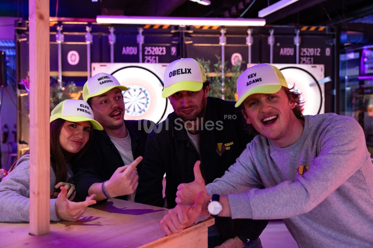 Quatre personnes souriantes portant des casquettes personnalisées posent devant un jeu de fléchettes lumineux.