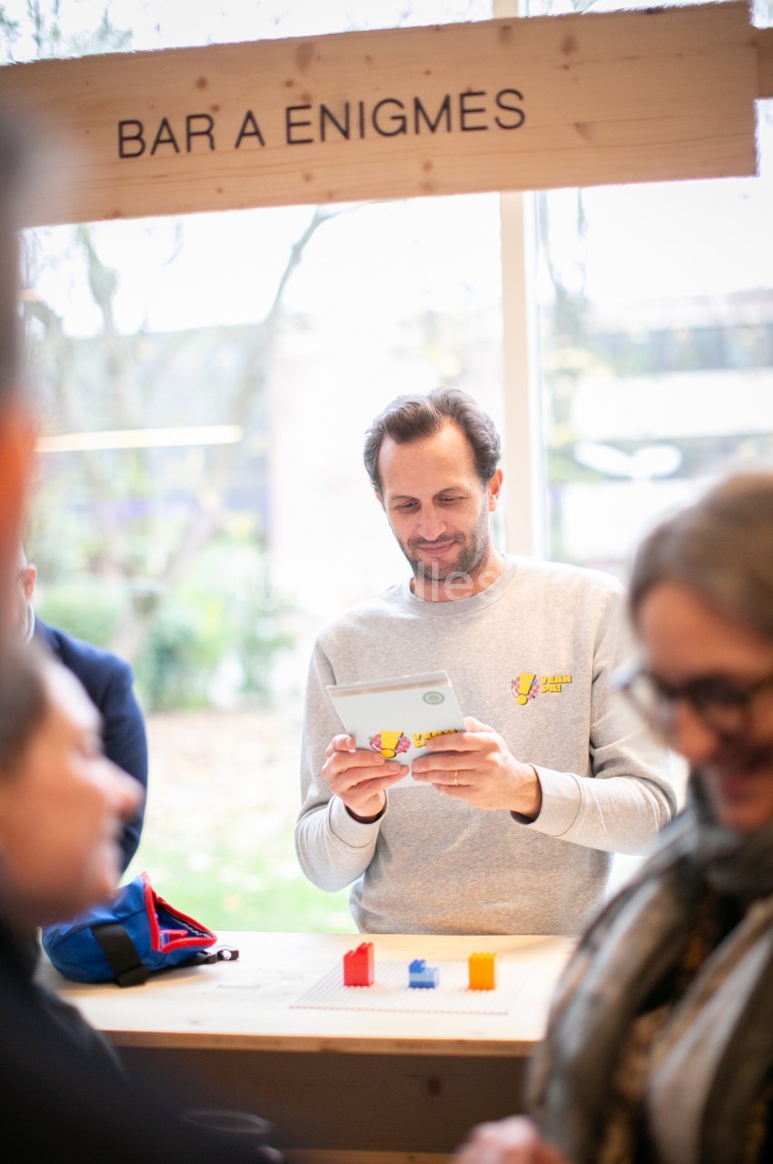 Homme lisant une tablette devant un stand de jeu intitulé ’Bar à énigmes’, avec des blocs colorés sur la table.