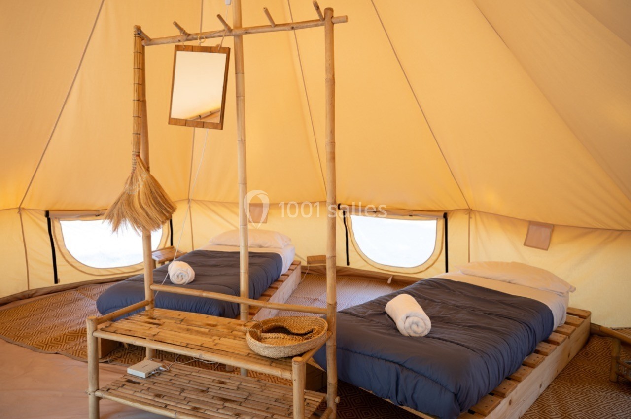 Intérieur d'une tente aménagée avec deux lits simples, linge de lit bleu, miroir suspendu et mobilier en bambou.