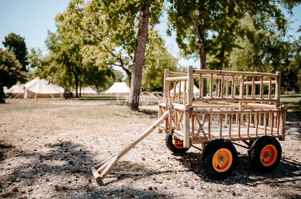 Chariot en bois avec roues orange, posé sur un sol gravillonné dans un espace extérieur arboré avec des tentes en arrière…