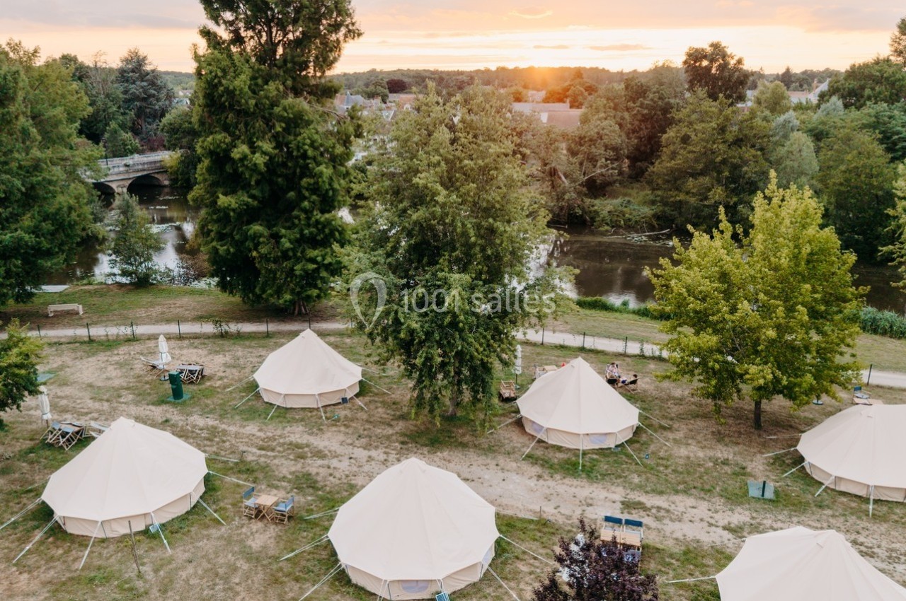 Tentes blanches installées dans un espace naturel près d'une rivière, entourées d'arbres et d'un pont au coucher du soleil.