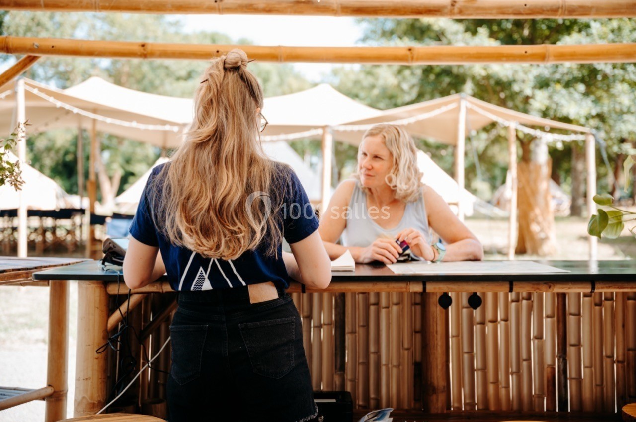 Une femme discute avec une employée derrière un comptoir en bois dans un cadre extérieur avec des tentes en arrière-plan.