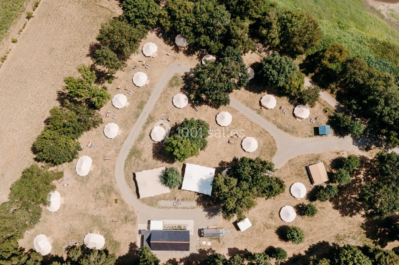 Vue aérienne d'un camping avec des tentes blanches disposées autour de chemins sinueux au milieu d'arbres et de prairies.