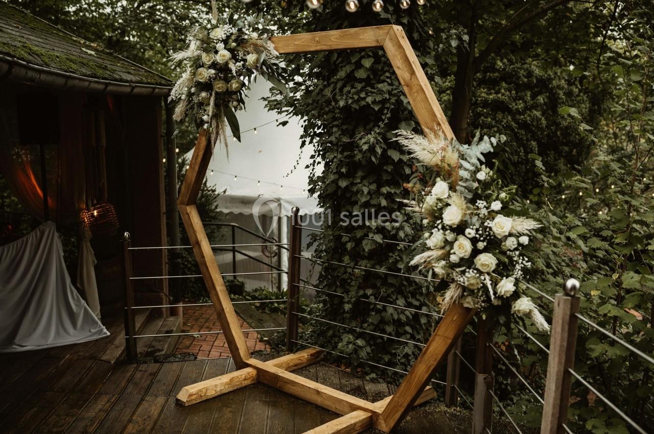 Arche hexagonale en bois décorée de fleurs blanches et feuillages, installée dans un cadre extérieur verdoyant.
