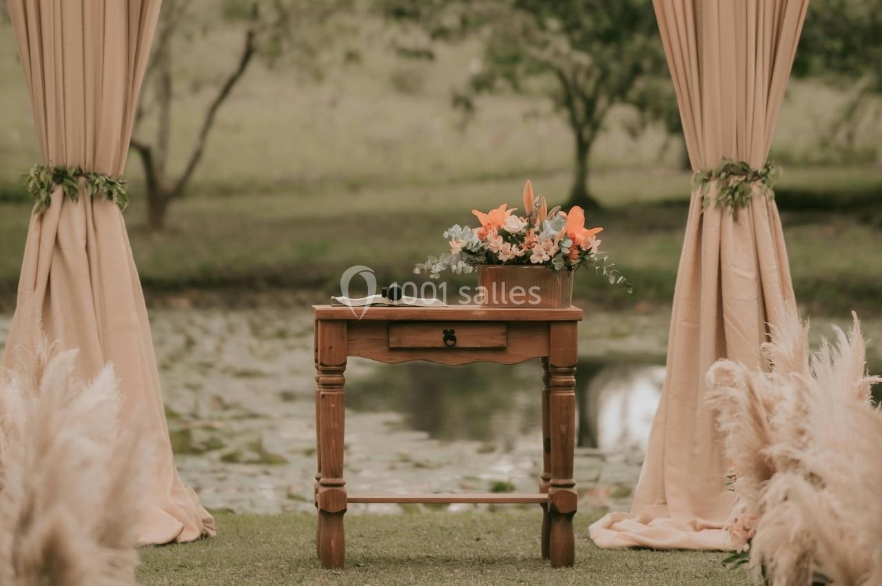 Arche de mariage décorée de fleurs et de tissus beige, avec une table en bois, dans un cadre naturel verdoyant.