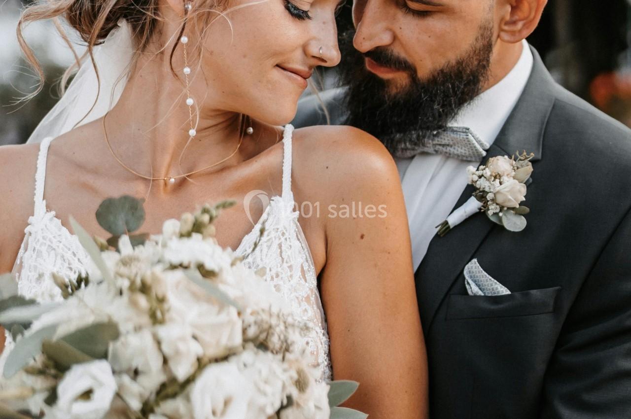 Un couple en tenue de mariage, la mariée tenant un bouquet de fleurs blanches, dans un cadre extérieur verdoyant.