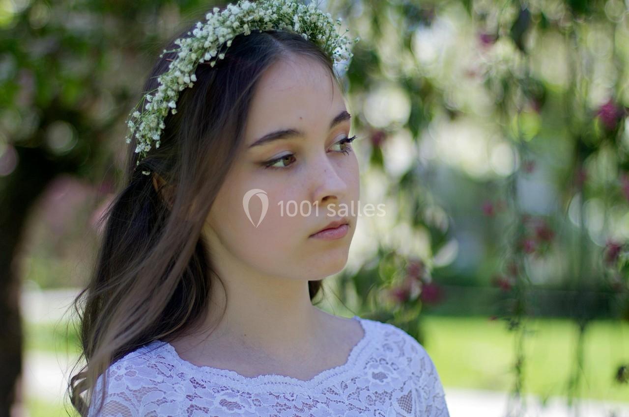 Jeune femme portant une couronne de fleurs blanches, vêtue de dentelle blanche, dans un jardin verdoyant.