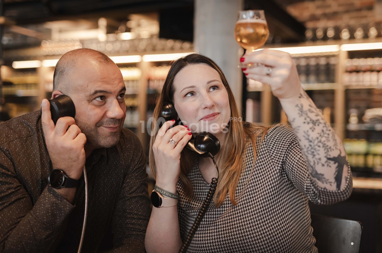 Un homme et une femme tenant des combinés téléphoniques, la femme examine un verre de boisson dorée.