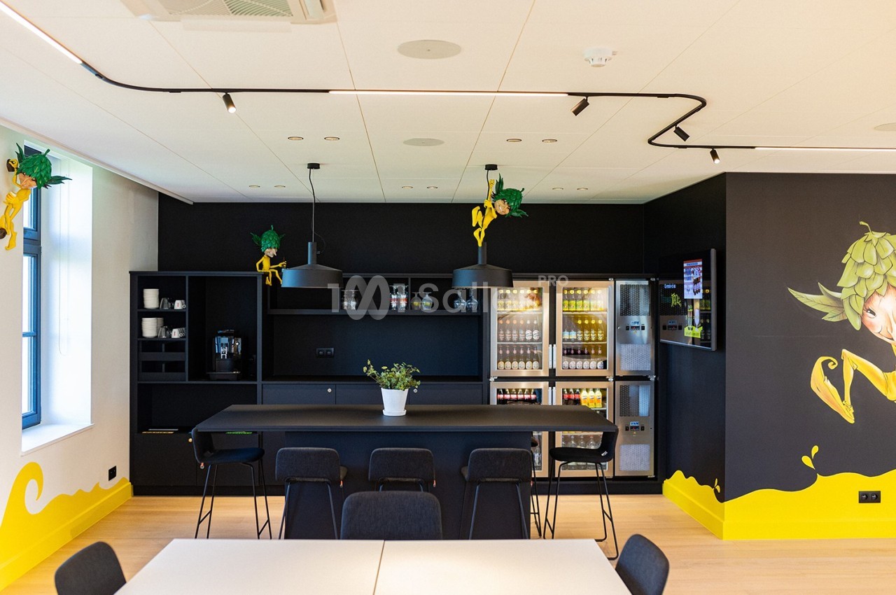Cuisine moderne avec comptoir noir, chaises, réfrigérateurs vitrés, machines à café et décoration murale colorée.