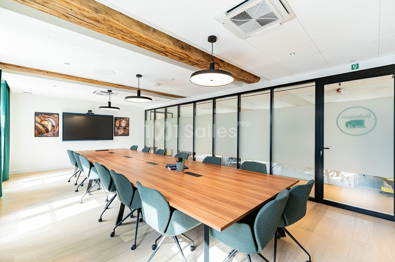 Salle de réunion lumineuse avec une grande table en bois, des chaises vertes et des cloisons vitrées.