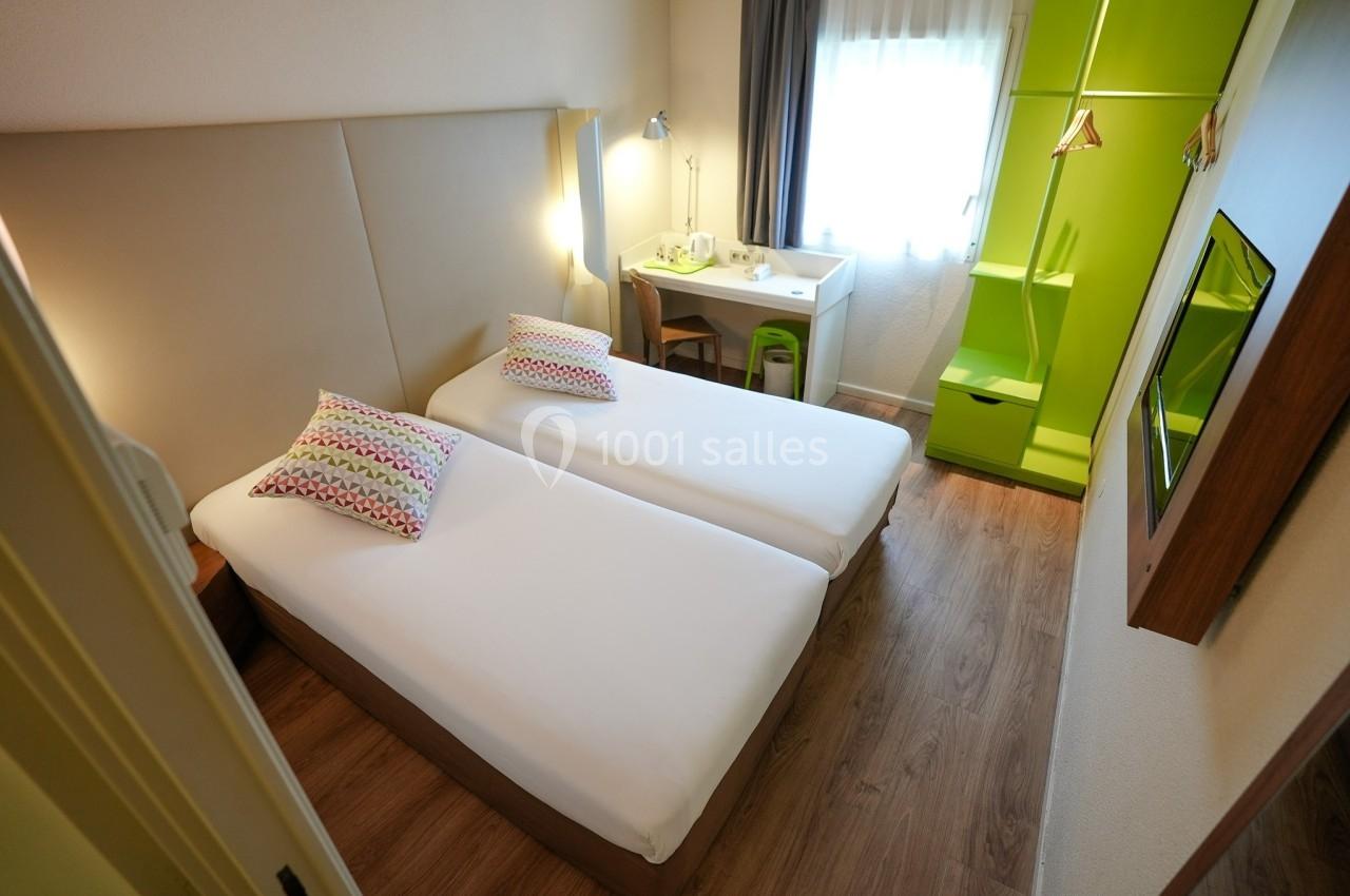 Chambre d'hôtel avec deux lits simples, bureau, chaise, armoire verte et fenêtre laissant entrer la lumière naturelle.
