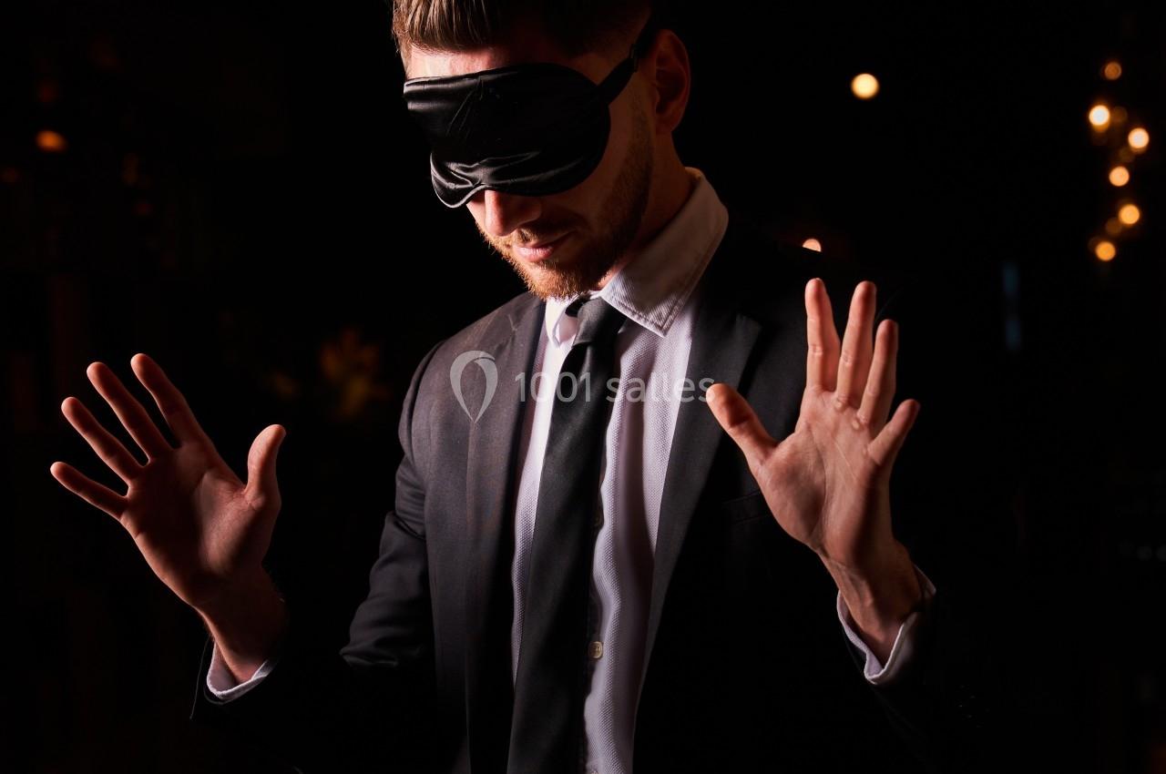 Un homme en costume, les yeux bandés, lève les mains dans un environnement sombre avec des lumières en arrière-plan.