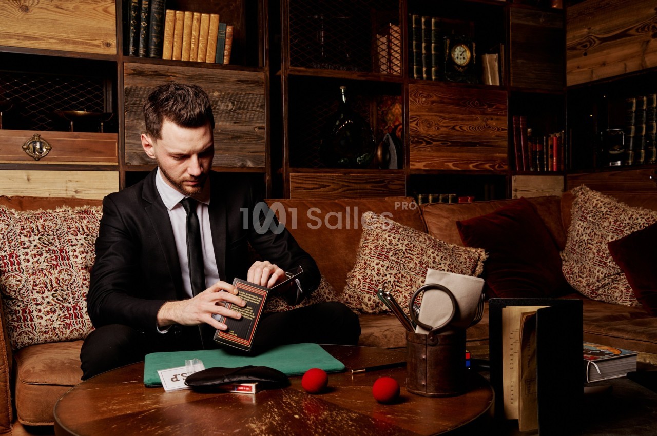 Un homme en costume assis dans un salon cosy, manipulant un livre entouré de documents et d'objets divers.