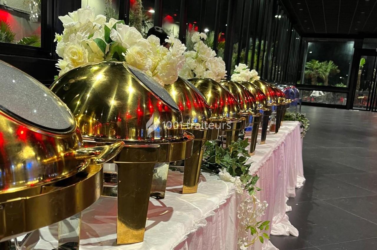 Rangée de plats en métal doré avec couvercles bombés, décorés de fleurs blanches, sur une table drapée.