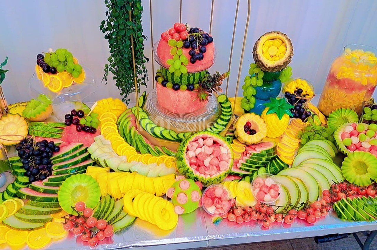Table garnie de fruits variés joliment découpés, incluant pastèques, ananas, raisins et melons, disposés en motifs…