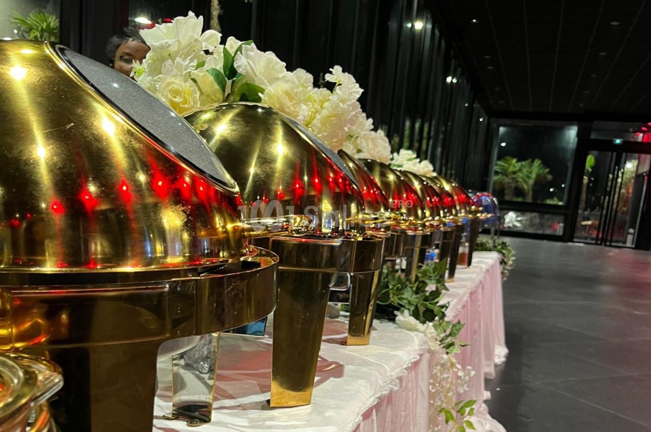 Rangée de plats en métal doré sur une table décorée de fleurs blanches dans une salle sombre.