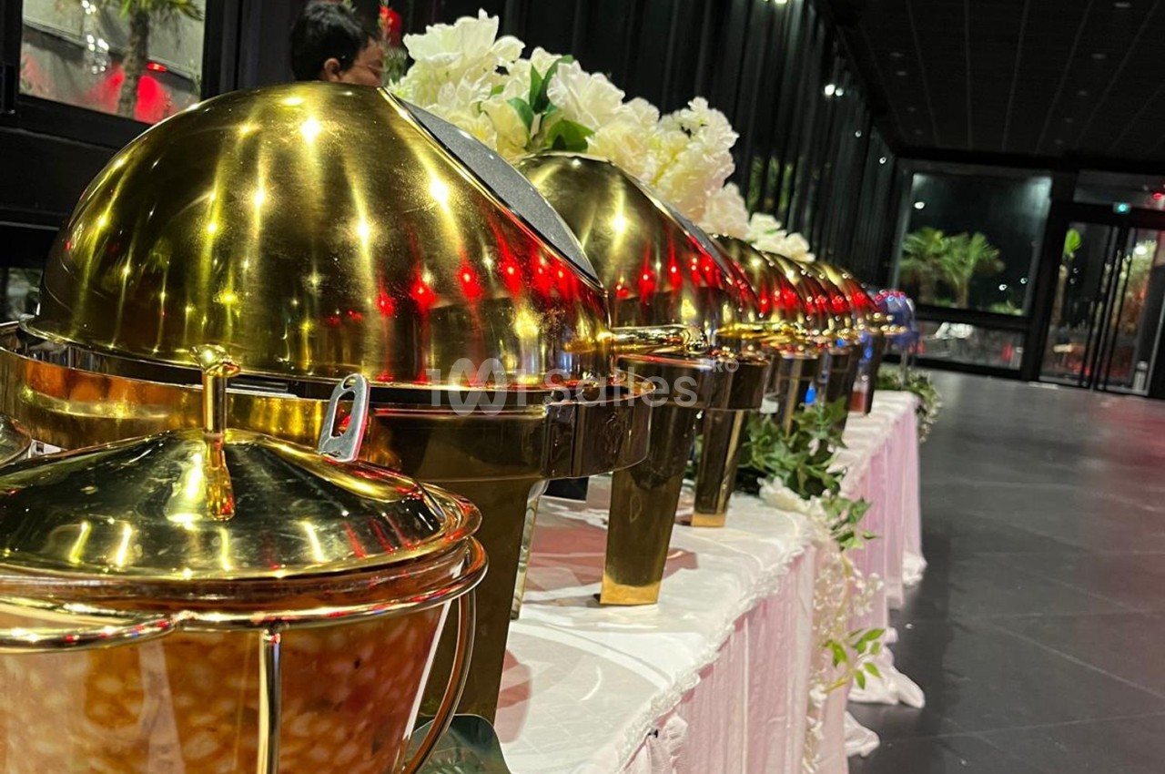 Rangée de plats en métal doré sur une table, dans une salle avec des baies vitrées et une décoration florale.