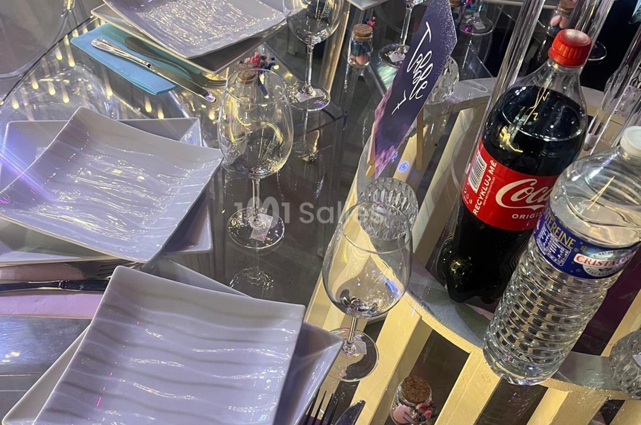 Table dressée avec des assiettes blanches, verres, bouteilles d'eau et de soda sur une surface réfléchissante.