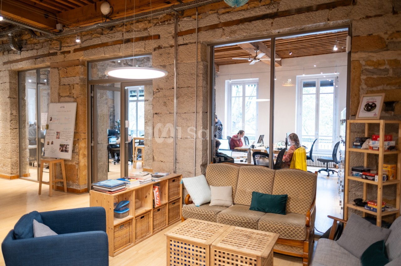 Espace de coworking lumineux avec canapés, étagères et vue sur une salle de travail vitrée en arrière-plan.