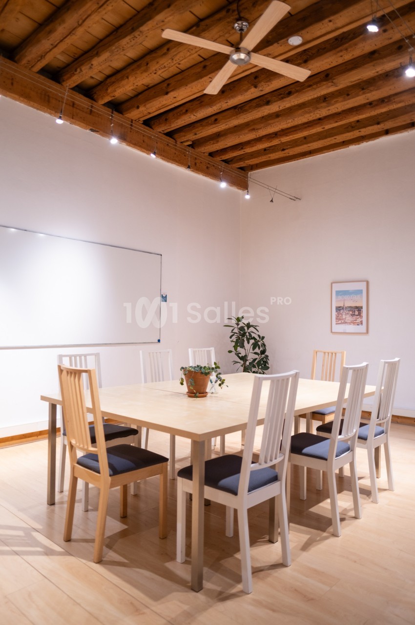 Salle de réunion lumineuse avec table en bois, chaises blanches, tableau blanc, plante et plafond en bois avec ventilateur.