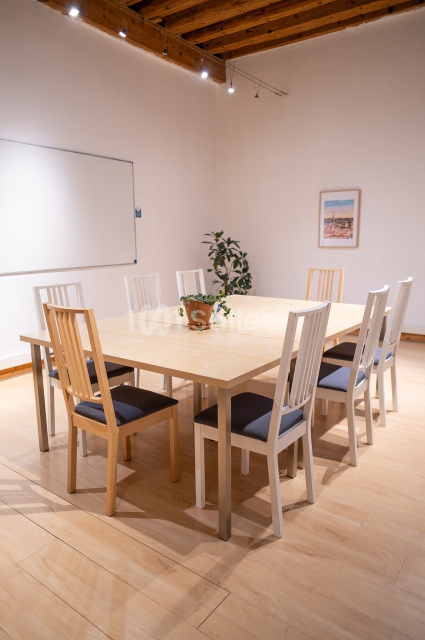 Salle de réunion lumineuse avec une grande table en bois entourée de chaises, un tableau blanc et une plante décorative.