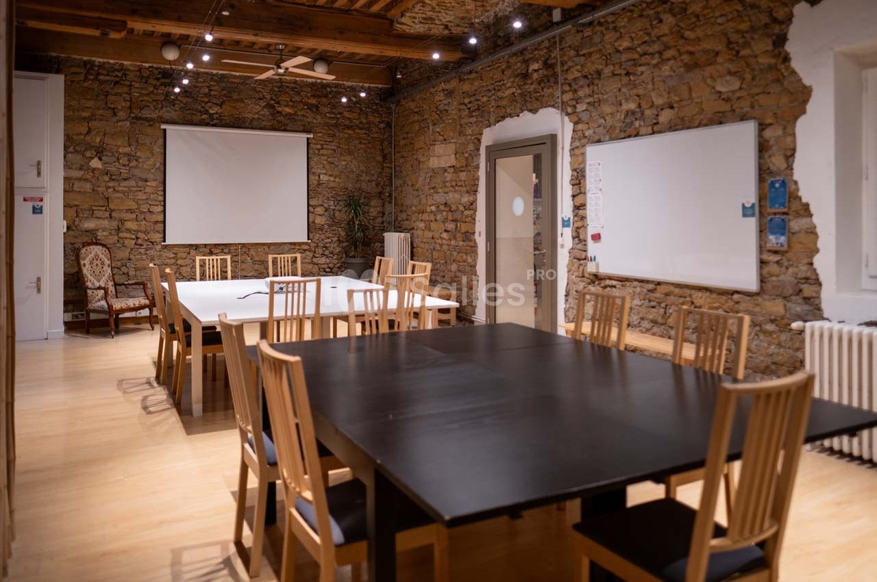 Salle de réunion avec murs en pierre, tables en bois, chaises, tableau blanc et écran de projection.