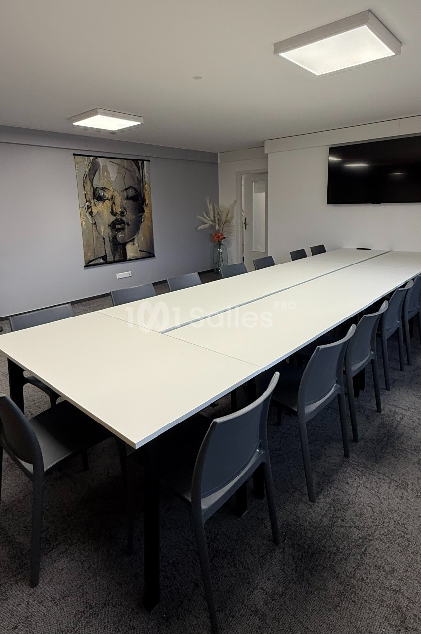 Salle de réunion moderne avec une grande table blanche, des chaises grises, un écran mural et une décoration minimaliste.
