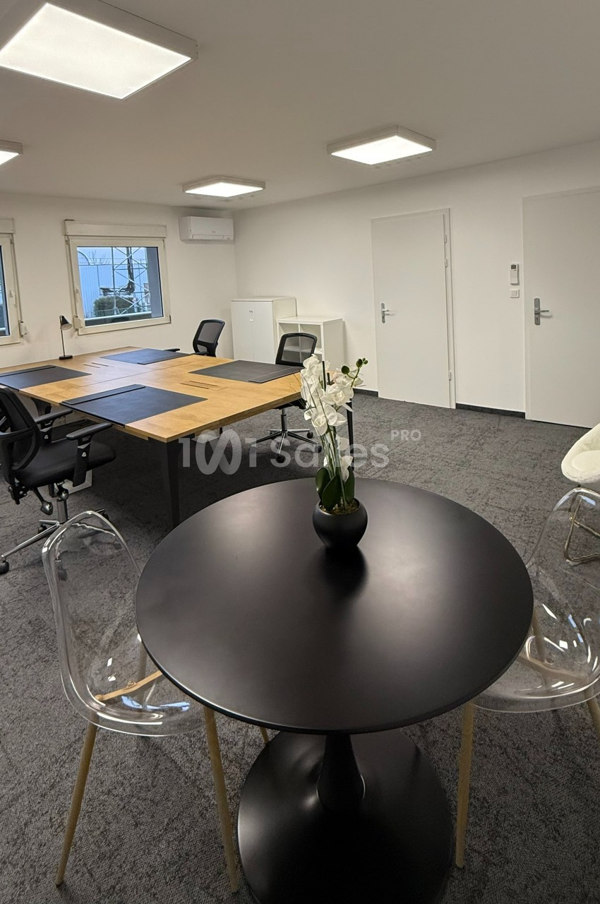 Salle de réunion moderne avec table ronde, chaises transparentes, bureau partagé et éclairage au plafond.