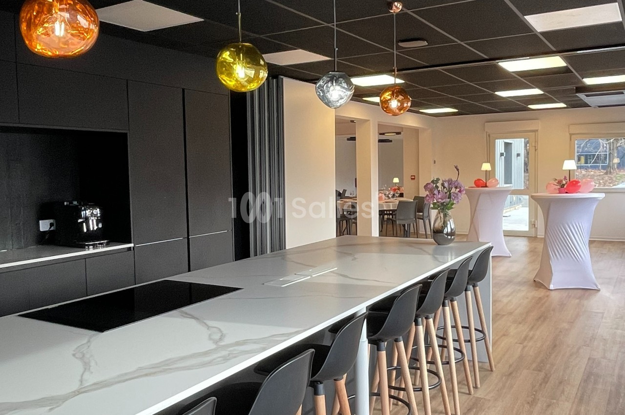 Cuisine moderne avec îlot central, tabourets noirs, suspensions colorées et vue sur une salle aménagée pour un événement.