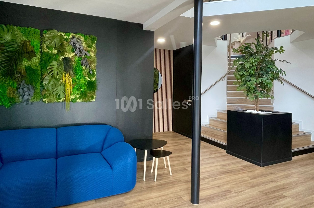 Espace intérieur moderne avec canapé bleu, mur végétalisé, escalier en bois et plantes décoratives.