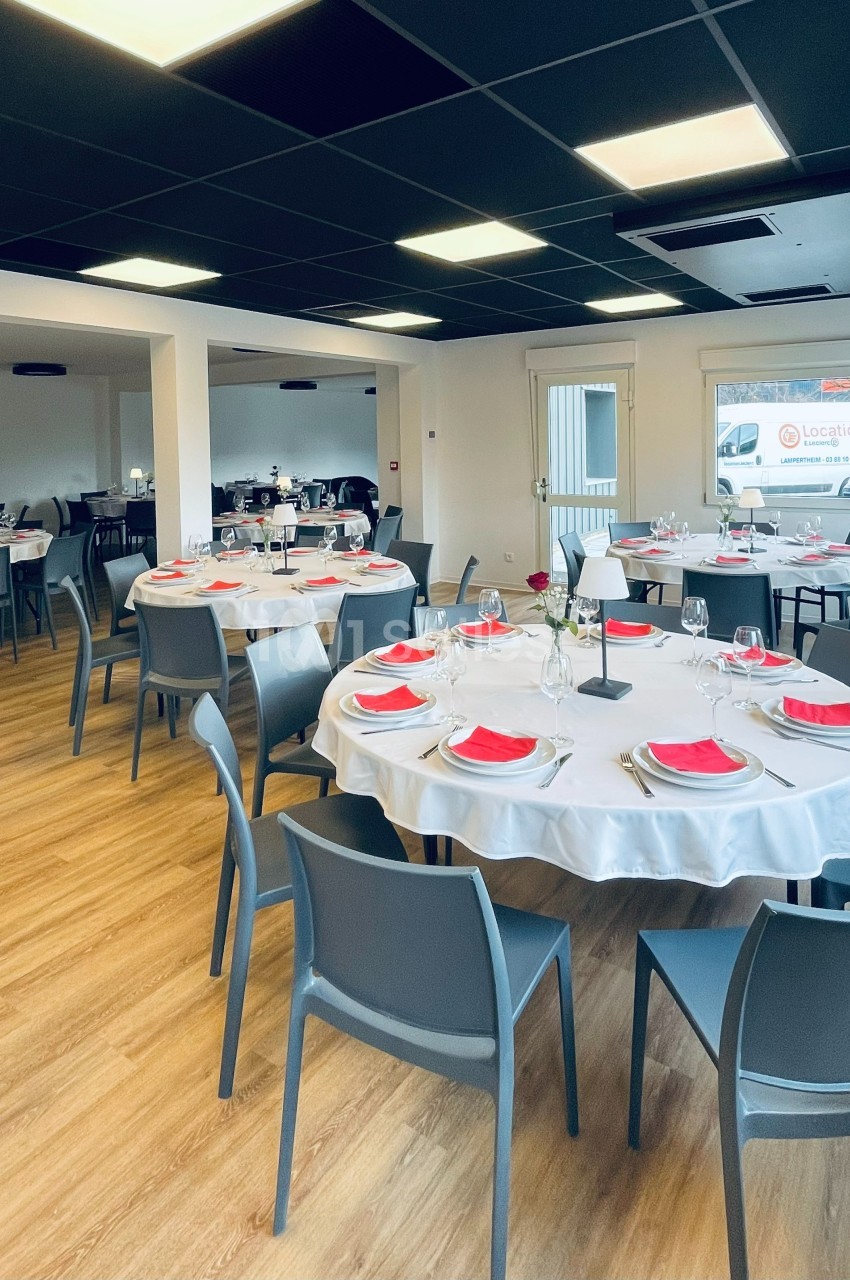 Salle de réception avec tables rondes dressées, nappes blanches, serviettes rouges et chaises grises sur parquet clair.