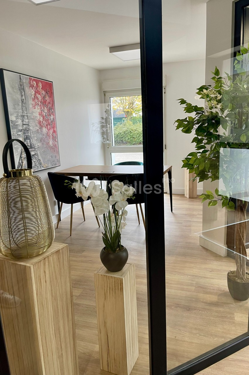 Salle lumineuse avec table en bois, chaises noires, plantes décoratives et tableau mural représentant la Tour Eiffel.
