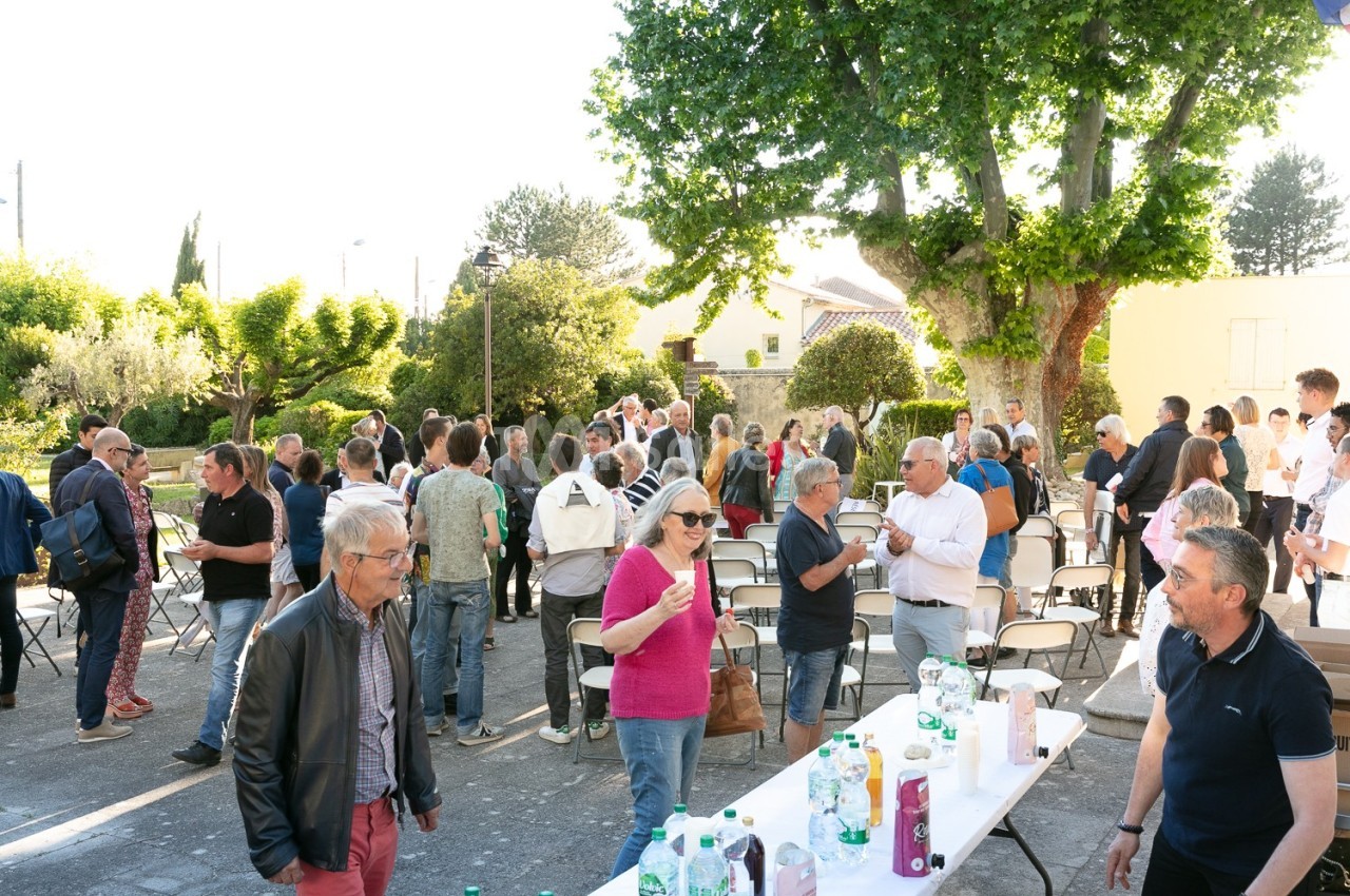 Groupe de personnes rassemblées en extérieur lors d'un événement, discutant près de tables avec boissons et verres.